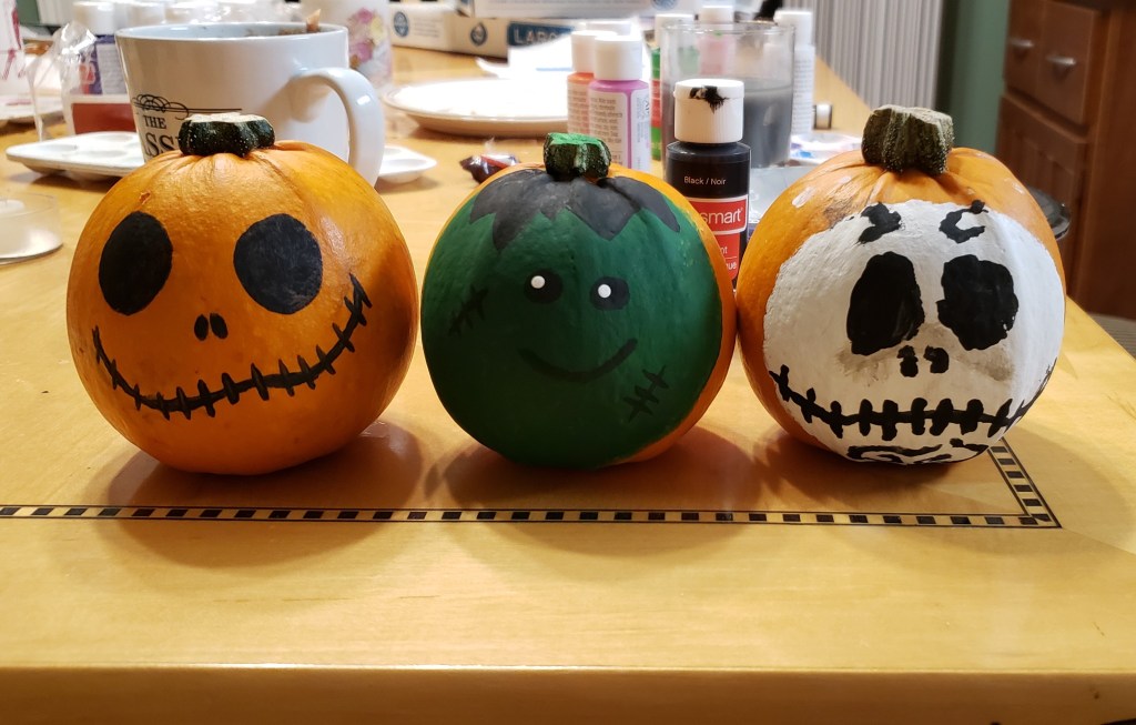 Three decorated mini-pumpkins with different faces sit on a light wooden table surrounded by painting supplies.