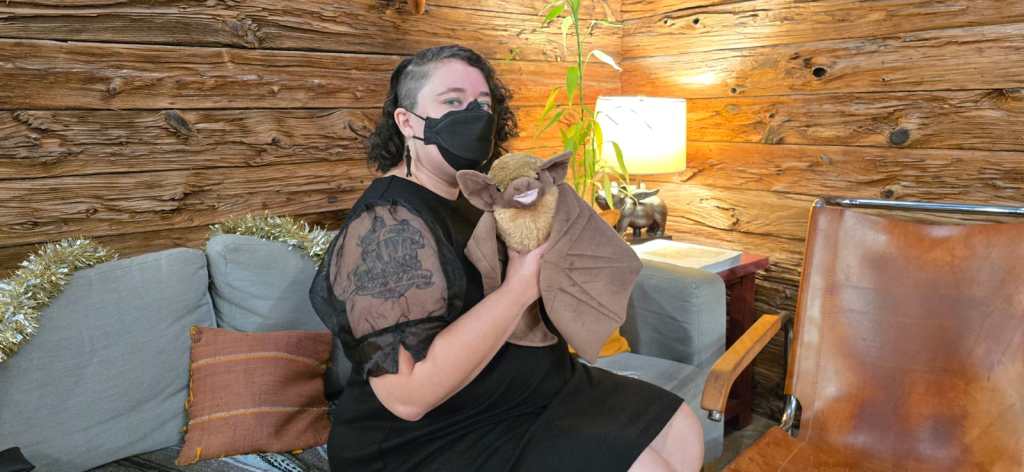 Leticia, a light skinned woman with dark curly hair with one side shaved sits on a gray sofa. She wears a black dress with puffy see through sleeves, black earrings and a black respirator mask. She holds a brown stuffed animal bat. There is wood paneling, a flying pig lamp and a plant in the background of the photo.