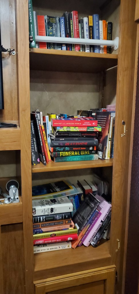 A brown RV built-in bookshelf full of books stacked and standing on three shelves. 