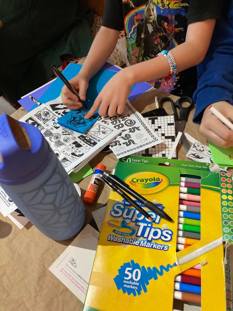A teen working on a zine at a wooden table filled with papers, markers and other materials.