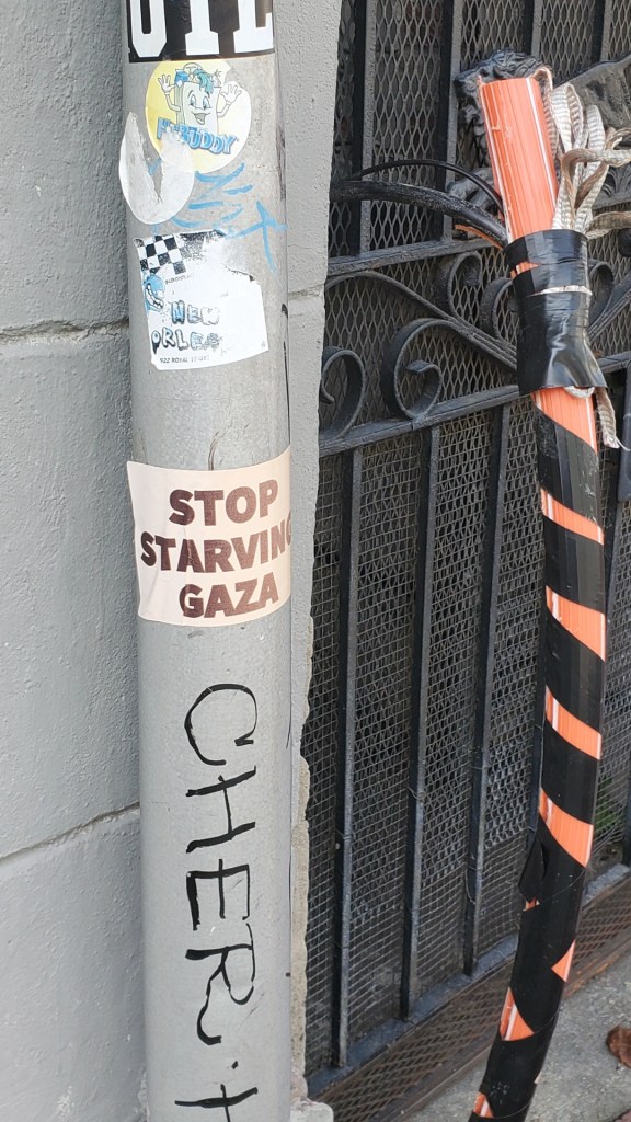 A metal light pole with stickers on it, one of which is square beige sticker with bold red text that says Stop Starving Gaza. 