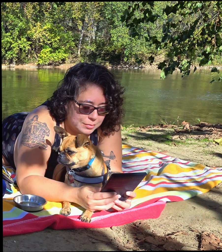 Leticia, a fat light skinned person with curly brown hair and black glasses lies on her stomach on a striped beach towel reading a book on her eReader with her pug mix, Tajin, sitting in between her arms. They lie under a tree by a river.  