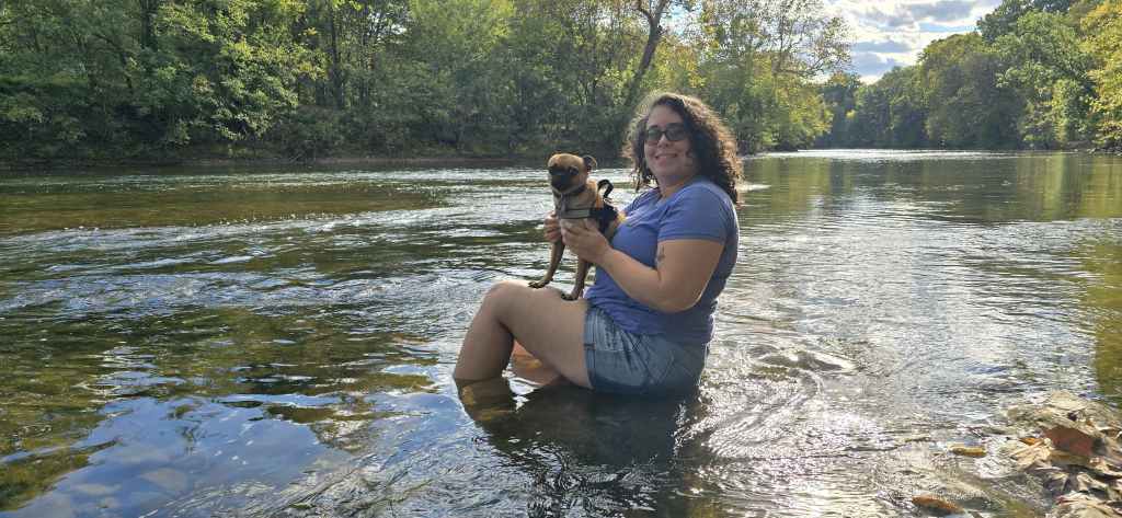 Leticia, a light skinned person with dark brown curly hair and black glasses wearing a sky-blue tshirt and jean shorts, sits on a rock on the shallow Shenandoah River with Tajin, her pug mix on her lap. 