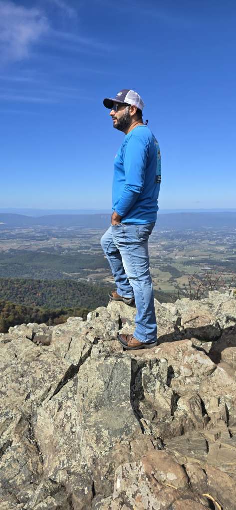 Ramiro, a brown-skinned tall man wearing a baseball cap, black sunglasses, a blue long sleeved shirt, blue jeans and brown hiking boots, poses with his foot on a rock outcropping overlooking the Shenandoah Valley. 