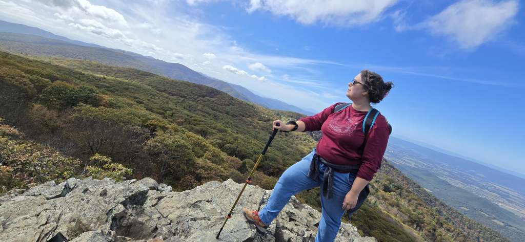 Leticia, a light skinned person with dark brown curly hair and black glasses wearing a long sleeved maroon red shirt, blue jeans and a black backpack, poses with her foot on a rock outcropping overlooking the Shenandoah Valley. 