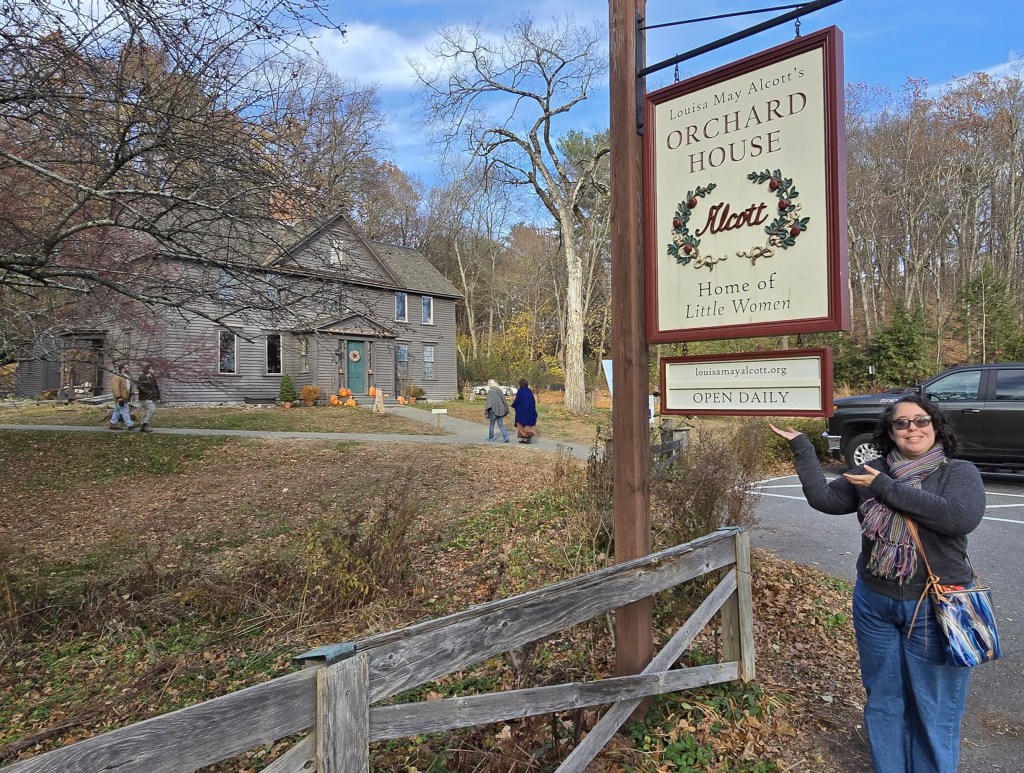 Leticia, a light skinned fat woman with curly dark brown hair, black glasses, a black sweater, jeans and a multicolored scarf and blue purse, poses in front of the wooden sign that says, Louisa May Alcott's Orchard House, home of Little Women, open daily. Behind her is Orchard House and people walking in the gardens around it.