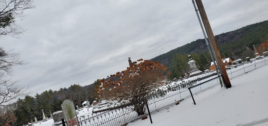 A small snow covered cemetery nestled in the green Vermont mountains with a black gate encircling it. 