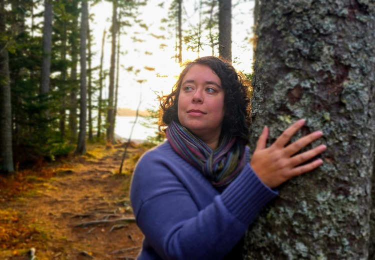 Leticia, a fat light skinned woman with dark brown curly hair and green eyes is looking into the distance with her hand on a mossy tree. She is wearing a blue sweater and a multicolored scarf. She stands against the tree in a wooded area on the coast of Maine. 