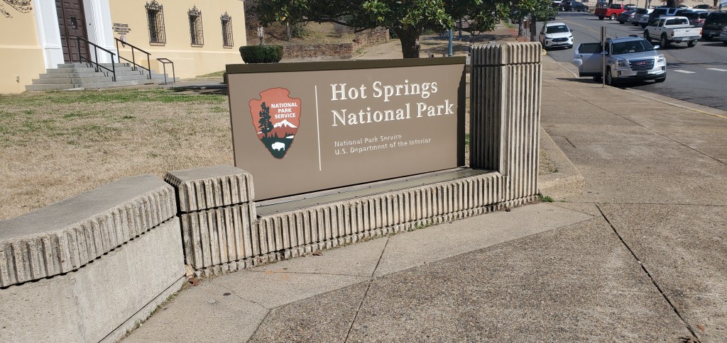 The brownish gray sign for the Hot Springs National Park in Arkansas placed on a sidewalk. 