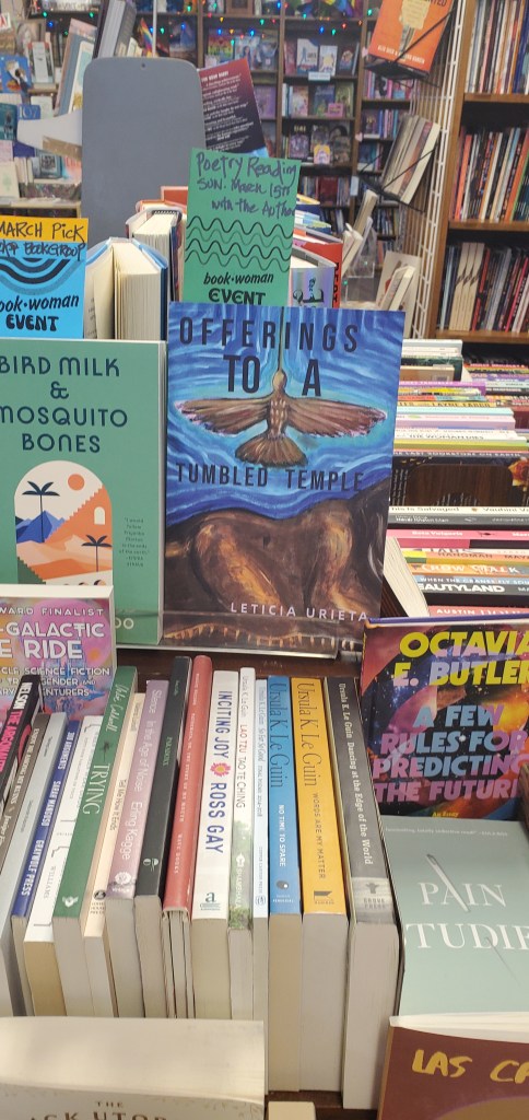 A copy of Leticia's book, Offerings to a Tumbled Temple, displayed on a table stacked with an array of other books, with bookshelves of Bookwoman Bookstore and twinkle lights in the background. The green note taped to Leticia's book says, "Poetry Readng Sunday March 15th with the author, bookwoman event." 