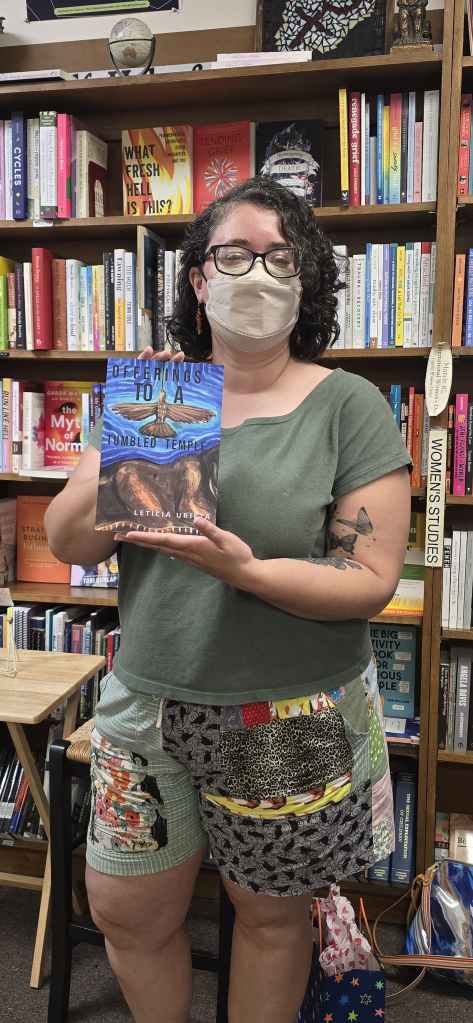 Leticia, a light skinned person with black short curly hair wearing black glasses, carnelian earrings, a pastel beige Kn95 mask, a forest green shirt and patched shorts, stands in front of bookcases in a bookstore holding up her book, Offerings to a Tumbled Temple.