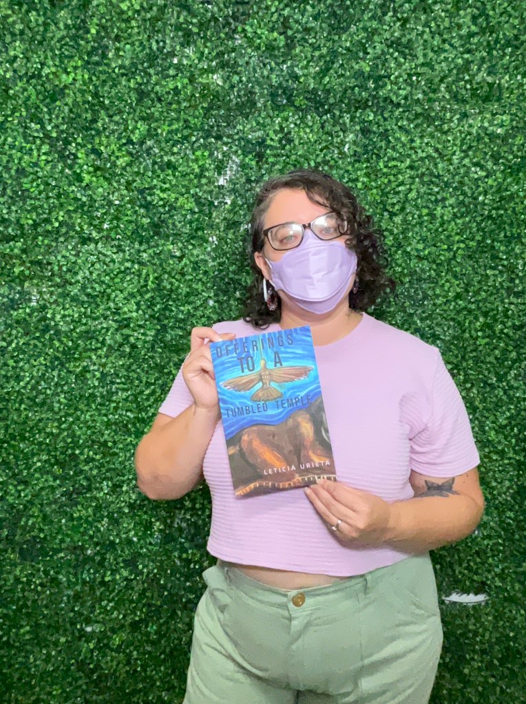 Leticia, a light skinned person with black curly hair, wearing black glasses, earrings, a lavender shirt and Kn95 mask and sage green trousers, stands in front of a green grass photo booth holding her book, OfferingstoaTumbled Temple, to the camera.