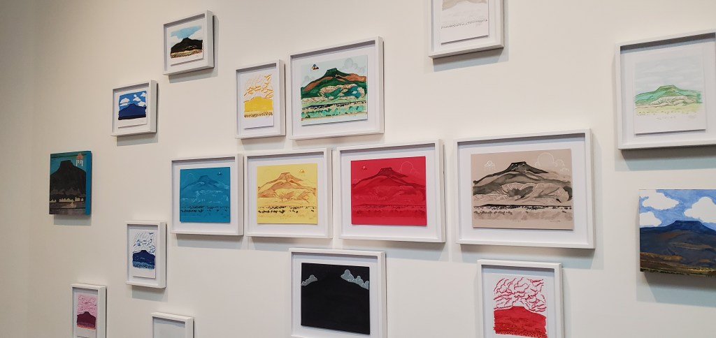 A series of illustrations and paintings in a variety of colors and materals in white picture frames on a white museum wall of tsi ping mountain or cerro pedernal near Santa Fe, New Mexico.