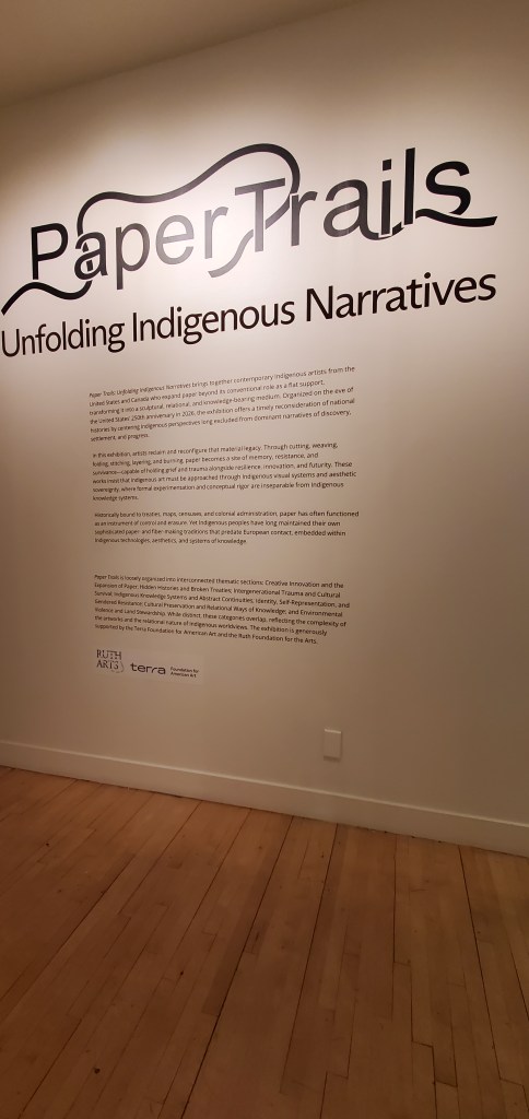 A beige wall with bold black text that says Paper Trails: Unfolding Indigenous Narratives, with a description of the exhibit in smaller black text.