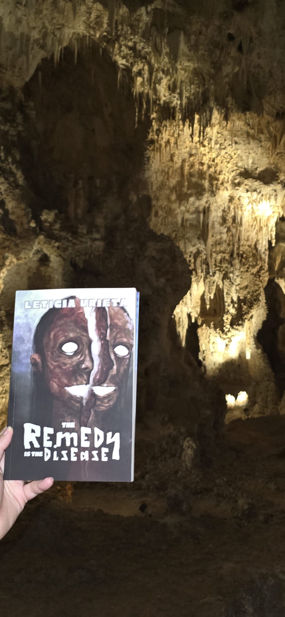 A copy of The Remedy is the Disease by Leticia Urieta held up by a white hand in a dimly lit black and beige cavern.
