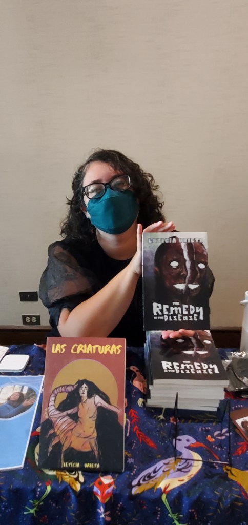 Leticia, a light skinned woman with black curly hair, black glasses, wearing a black dress and chartreuse mask holds her book, The Remedy is the Disease, up to the camera where she is sitting at a table.