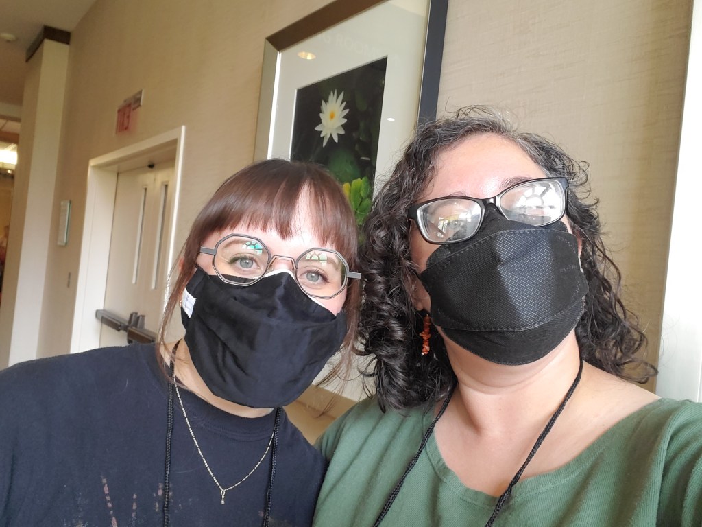 Michelle Delgado, a person with white skin and red hair wearing a black mask poses with Leticia Urieta, a weight skinned person with black curly hair, black glasses wearing a forest green shirt and black respirator mask.