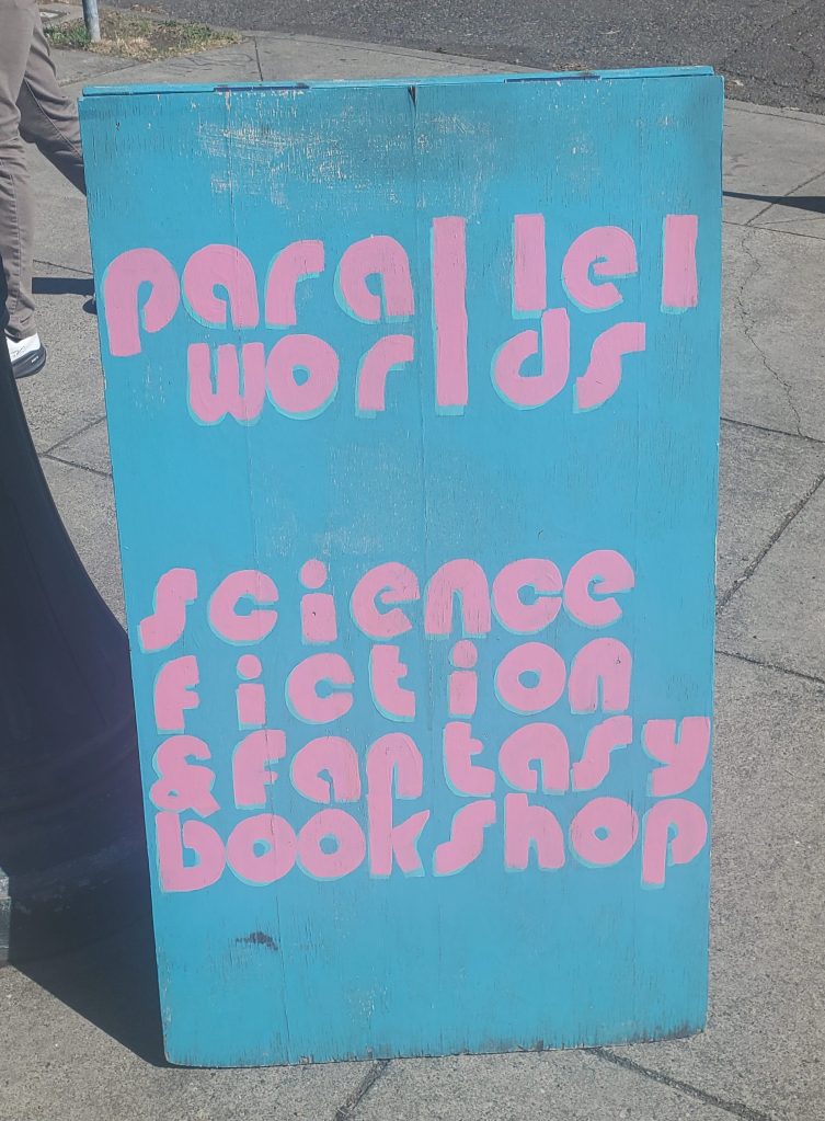 The sky blue and pink sandwich board sign for Parallel Worlds Science Fiction & Fantasy Bookshop in Portland, Oregon.