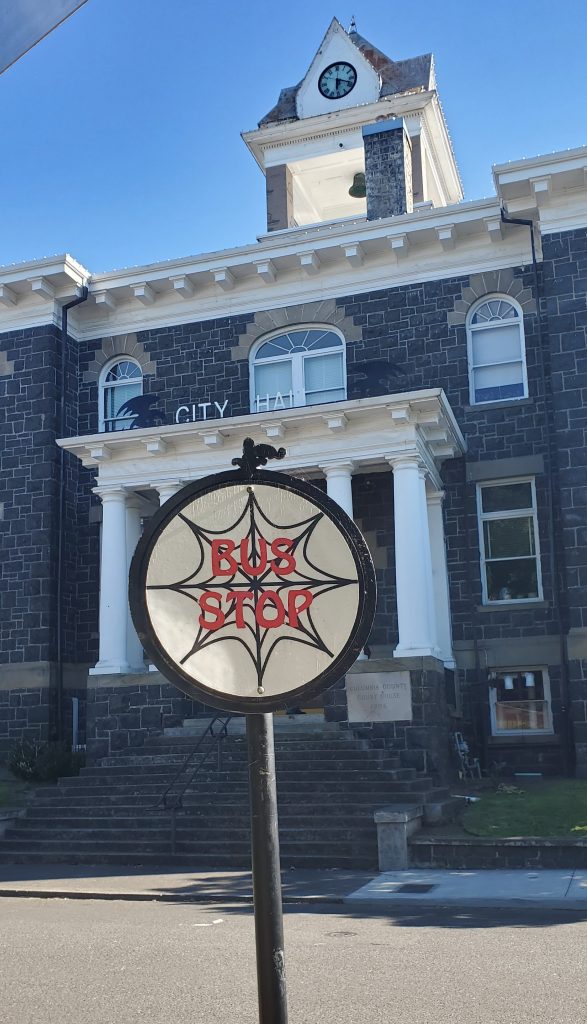 A black and white round sign with a spiderweb in the middle and the words Bus Stop on it in bold red letters stands in front of a gray brick City Hall building with white columns.