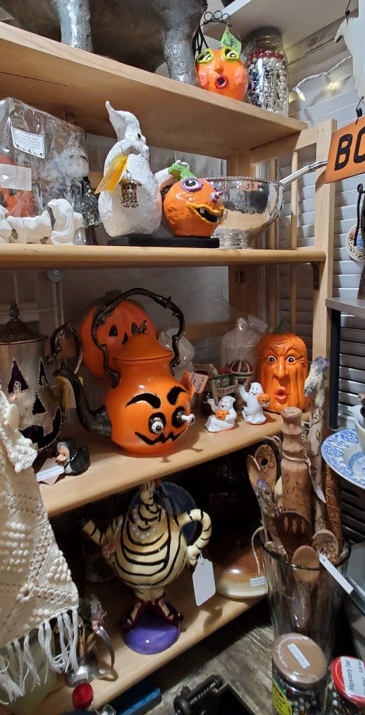 A shelf full of antique figurines, bowls and Halloween decorations, including an orange pumpkin copper teapot with white eyes sticking out.