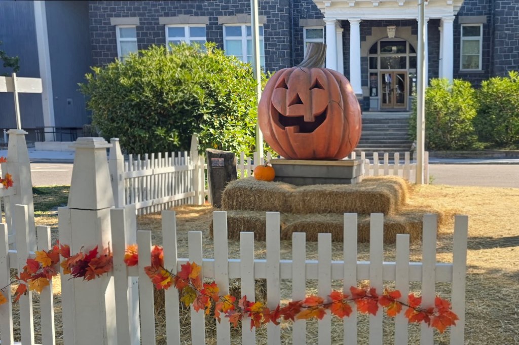The large orange Jack-o-lantern from Halloweentown sits on a stone pedestal in the town square in front of the gray brick City Hall building surrounded by hay bales and a white picked fence.