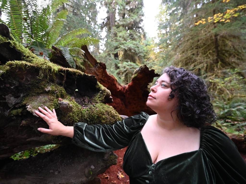 Leticia, a light-skinned woman with short curly dark brown hair wearing a forest green velvet long sleeved dress, stands in a forest with bright green ferns and trees, her hand resting on a mossy downed tree log as she looks into the distance.